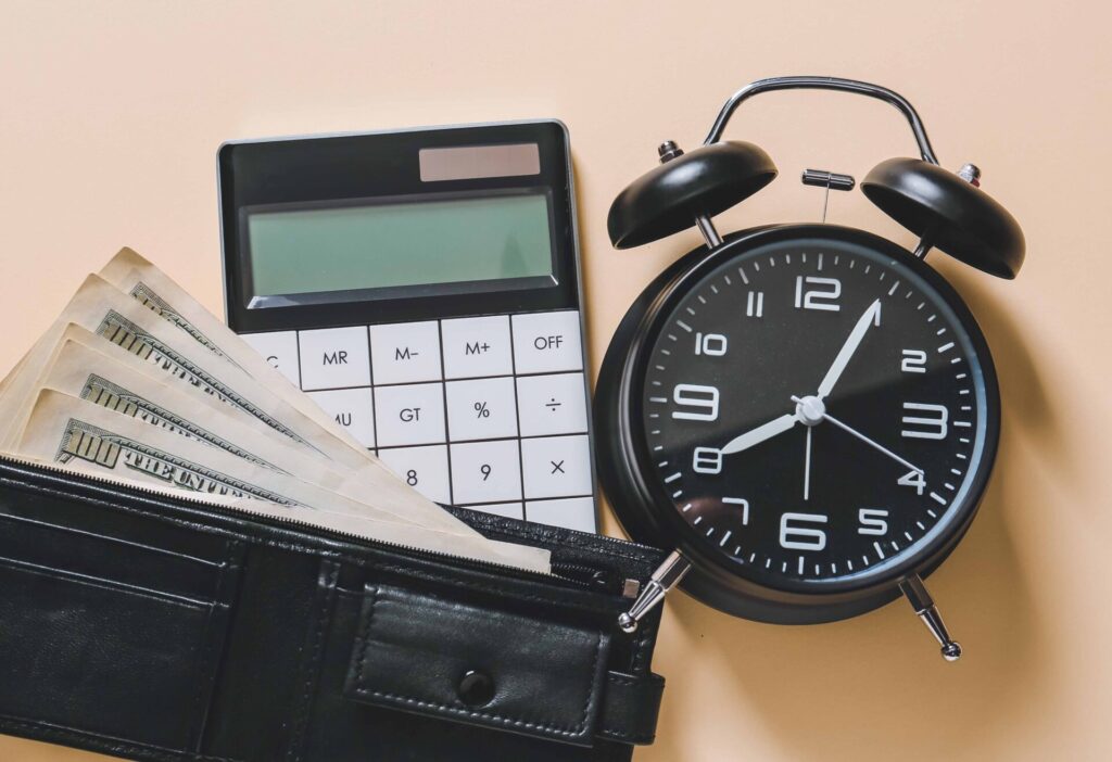 木のテーブルの上に黒い目覚まし時計、電卓、財布が置かれている