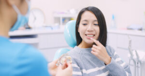 青いマスクとシャツを身につけた歯科医が、歯の模型を手に持って唇に人差し指をあてた患者に説明している