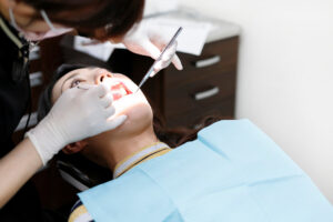 白いマスクと手袋をしている歯科医が、水色のエプロンをして仰向けになり口をあけている患者の口の中を確認している